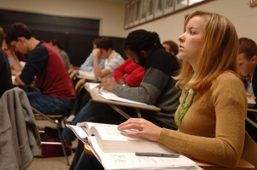 一群学生坐在课桌前，一个女人站在最前面望着前面.